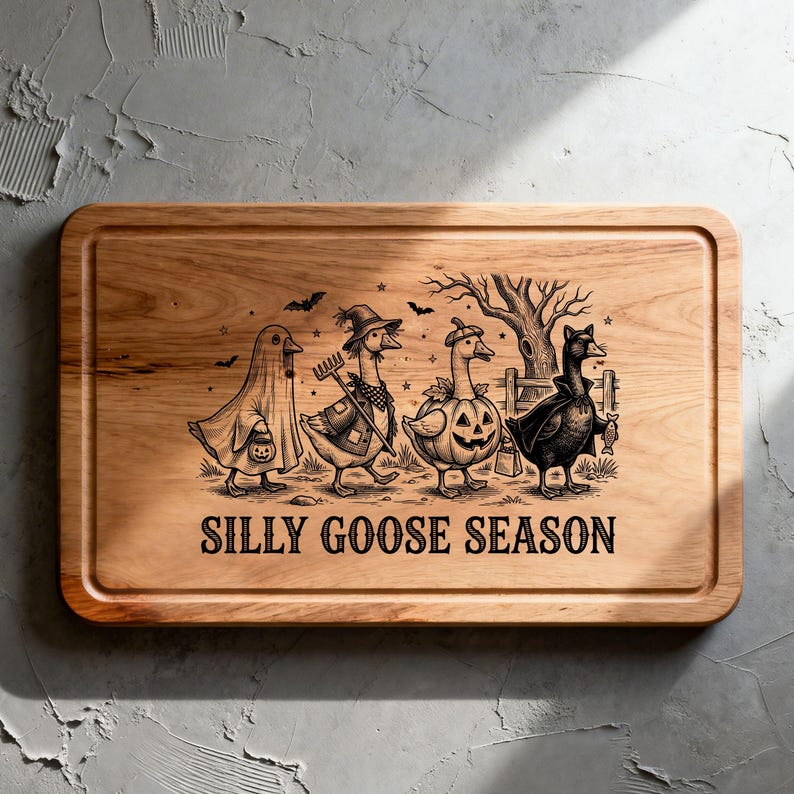 May include: A rectangular wooden cutting board with a grooved border featuring a black engraved illustration of four geese in Halloween costumes. The text SILLY GOOSE SEASON is printed below the geese, which include a ghost, a farmer, a pumpkin, and a vampire.