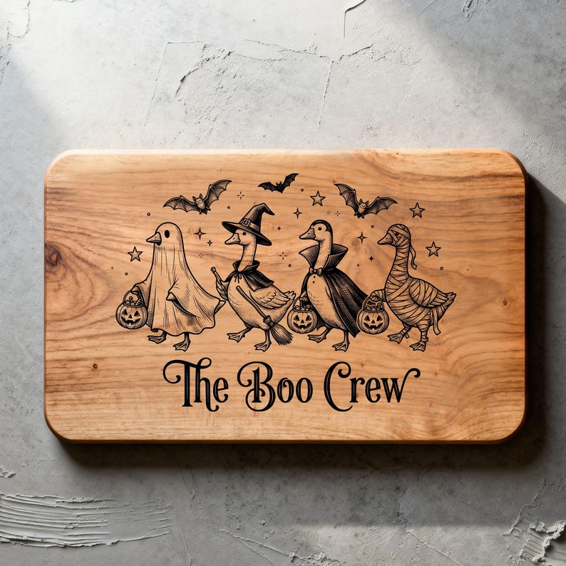 May include: A rectangular wooden cutting board featuring a black laser-engraved illustration of four geese dressed in Halloween costumes, including a ghost, a witch, a vampire, and a mummy, with the text "The Boo Crew" printed below them.
