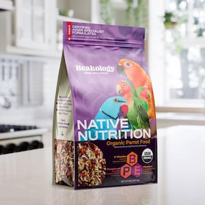 May include: A bag of Beakology Native Nutrition organic parrot food. The bag is purple and features colorful parrot illustrations. The text on the bag includes "Certified Avian Specialist Formulated" and "Organic Parrot Food". The net weight is 2 lbs.