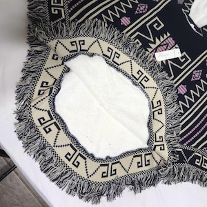 May include: A navy blue poncho with a geometric pattern in cream and lavender. The poncho has a cream-coloured border with black geometric shapes and fringe. The interior is white.