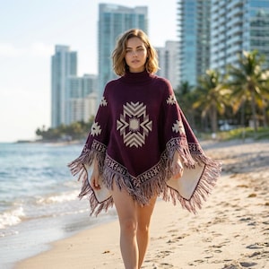 May include: A burgundy poncho with a geometric design in cream and tan, featuring a turtleneck and fringed edges. The poncho is worn on a beach with buildings in the background.