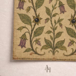 May include: A rectangular textile piece featuring a repeating botanical pattern on a beige linen-like background. The design displays vertical stems with green leaves, purple bell-shaped flowers, and dark circular accents. The letters AH are printed below.