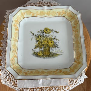 May include: Square, vintage porcelain serving dish with a yellow floral border and a central bouquet of yellow roses in a vase. The dish has a scalloped edge and gold trim. The dish is resting on a white lace doily.