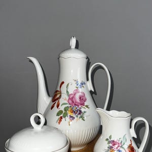 Puede incluir: Un juego de té de porcelana blanca con tetera, azucarero y jarra de crema. Cada pieza está adornada con un diseño floral en tonos rosa, morado y amarillo. La tetera tiene una forma alta y esbelta con un pico y asa curvos.