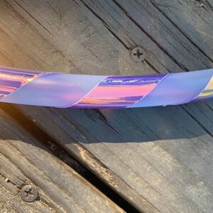 May include: A close-up of a section of a hula hoop with iridescent tape in shades of purple, pink, and yellow. The tape is wrapped around the hoop in a diagonal pattern. The background is a weathered wooden surface.
