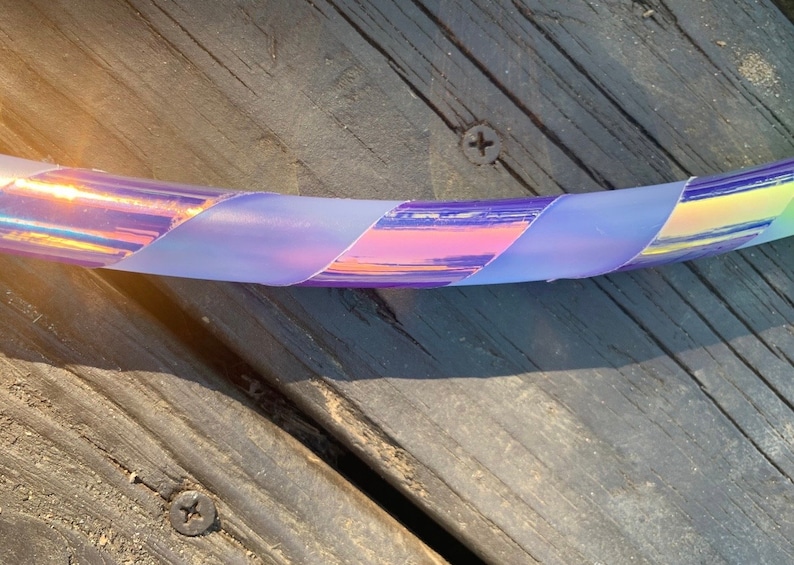 May include: A close-up of a section of a hula hoop with iridescent tape in shades of purple, pink, and yellow. The tape is wrapped around the hoop in a diagonal pattern. The background is a weathered wooden surface.