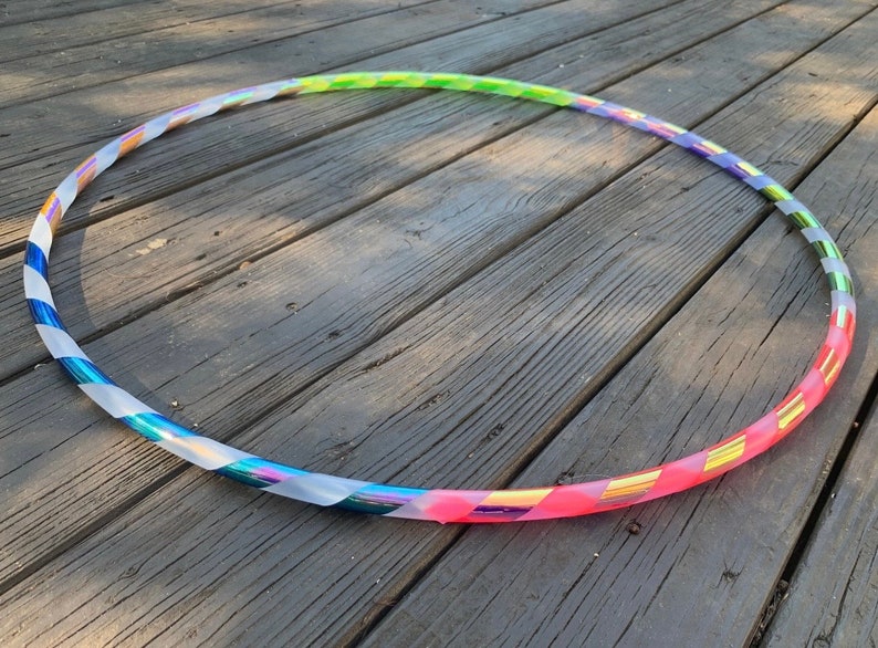 May include: A colorful hula hoop with a rainbow gradient design. The hoop features a clear, iridescent tape with diagonal stripes in shades of blue, green, yellow, and pink. The hoop is lying on a weathered wooden surface.