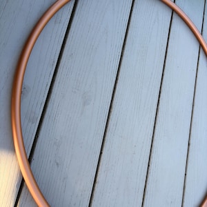May include: A large, circular hula hoop with a smooth, solid copper-colored finish. The hoop is constructed from a single continuous tube, forming a perfect ring shape. A portion of a second, smaller blue and green hula hoop is visible at the top.