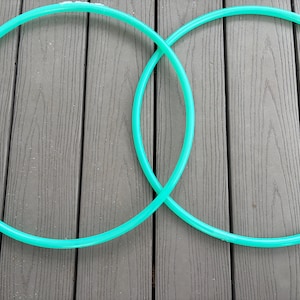Puede incluir: Dos aros de hula-hoop turquesa se cruzan sobre un fondo de madera gris. Los aros, de un verde vibrante, están hechos de un material liso y redondeado. Cada aro mide aproximadamente 90 cm de diámetro.