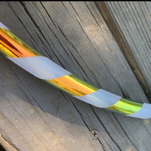 May include: A section of a colorful hula hoop. The hoop has a metallic gold and iridescent finish with sections of white tape wrapped around it. The hoop is resting on a weathered wooden surface.