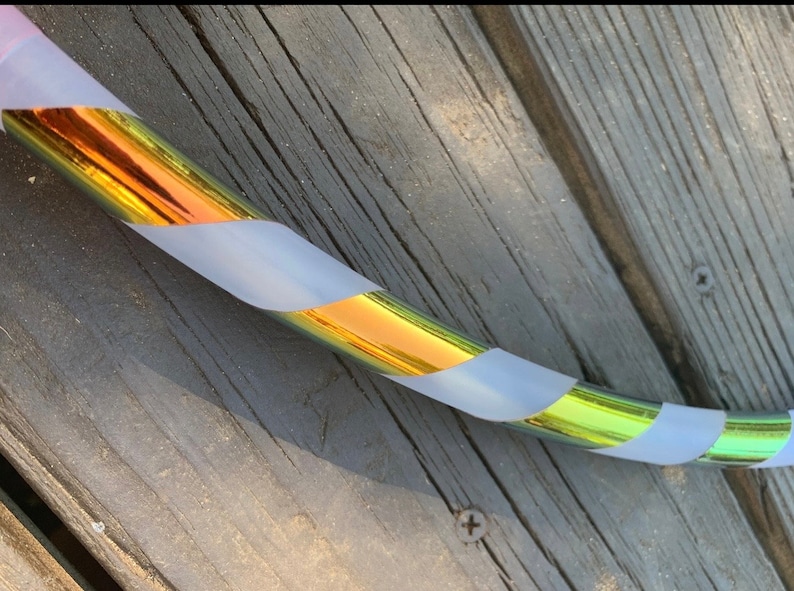 May include: A section of a colorful hula hoop. The hoop has a metallic gold and iridescent finish with sections of white tape wrapped around it. The hoop is resting on a weathered wooden surface.