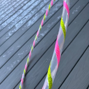 Puede incluir: Un aro de plástico transparente con cinta reflectante rosa y verde envuelta alrededor. El aro está sobre una superficie de madera gris.