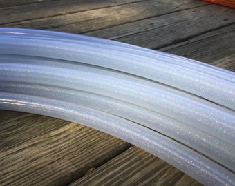 Puede incluir: Varios aros de hula de color blanco con purpurina iridiscente apilados uno encima del otro. Los aros están hechos de plástico transparente con un acabado brillante.