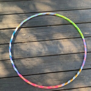 May include: A colorful hula hoop with a twisted design. The hoop transitions through shades of pink, purple, blue, green, and yellow. The hoop is lying on a weathered wooden surface.