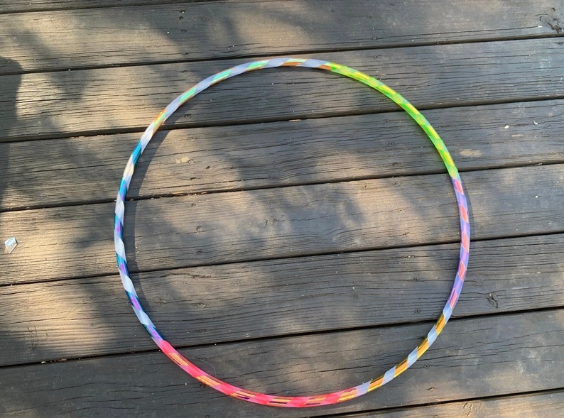 May include: A colorful hula hoop with a twisted design. The hoop transitions through shades of pink, purple, blue, green, and yellow. The hoop is lying on a weathered wooden surface.