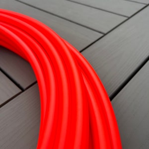 Puede incluir: Primer plano de un aro de hula naranja brillante. El aro está hecho de un material flexible y está enrollado sobre una superficie gris.