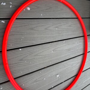 Puede incluir: Un aro de hula rojo brillante, de forma circular, descansa sobre una cubierta de madera gris. El aro parece estar hecho de un material liso y sólido. El fondo muestra una cubierta de madera con pequeños parches de nieve.