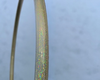 Può includere: Un cerchio dorato con glitter iridescente incorporato nella plastica. Il cerchio è parzialmente visibile su uno sfondo bianco.