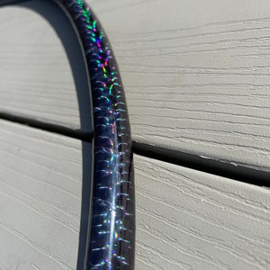 Puede incluir: Un aro negro con cinta hologr&aacute;fica iridiscente. La cinta tiene un patr&oacute;n de arco&iacute;ris con colores verde, azul, morado y rojo.