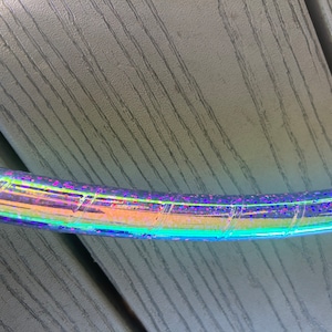 Puede incluir: Una sección de un hula hoop holográfico. El aro es iridiscente, reflejando un espectro de colores que incluyen azul, verde y rosa. La superficie tiene una textura brillante y está envuelta en un patrón en espiral.