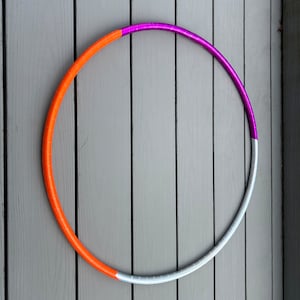 Puede incluir: Un aro de ejercicio colorido con secciones naranja, blanca y morada. El aro está acostado sobre una superficie de madera gris.