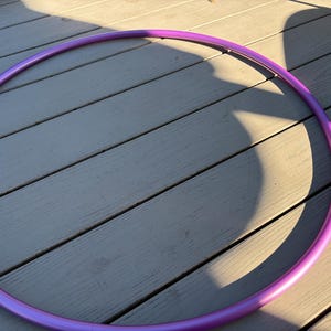Puede incluir: Un aro de hula hula circular con un acabado liso y de color morado sólido. El aro está construido a partir de una sola pieza continua de tubo, formando una forma de anillo perfecta. La superficie del material morado es uniforme y refleja.