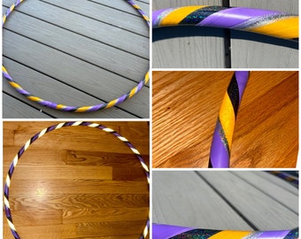 Könnte beinhalten: Ein lila, gelber und schwarzer gestreifter Hula-Hoop. Der Reifen ist aus Kunststoff und hat eine glänzende Oberfläche.