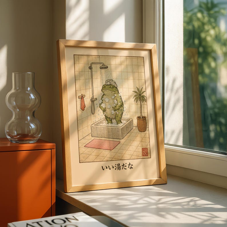 May include: A framed art print depicts a frog in a bath, with a showerhead and a small plant. The frog is covered in bubbles and wearing a towel on its head. The text "いい湯だな" is at the bottom of the print. The frame is made of light-colored wood.