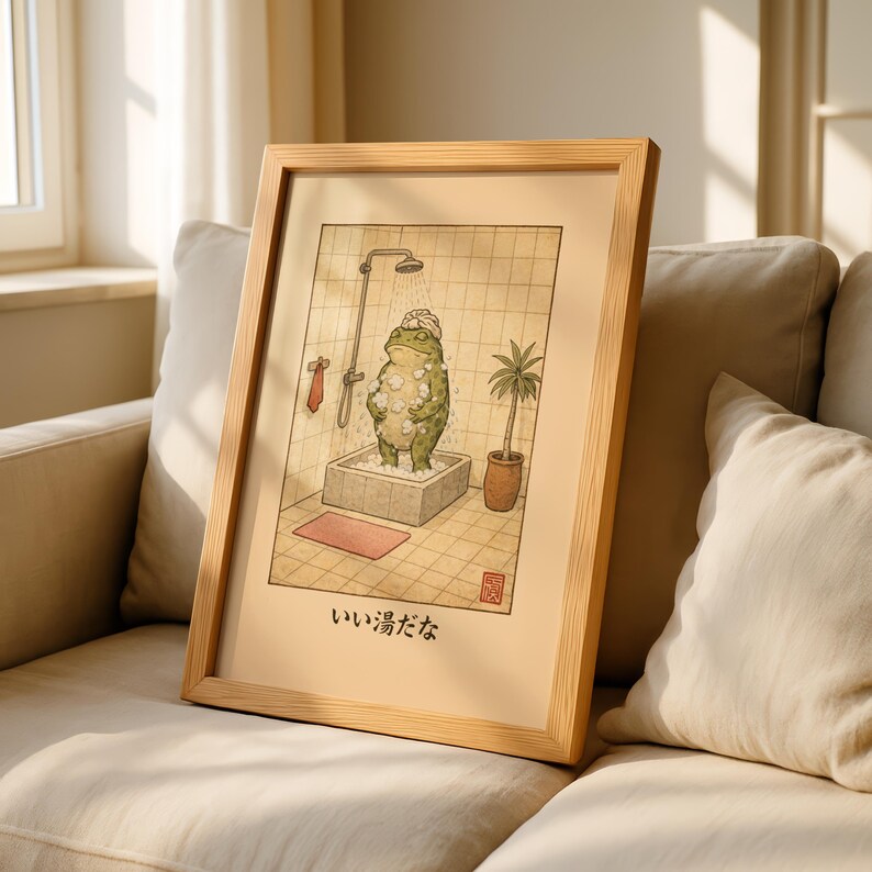 May include: Framed art print featuring a green frog taking a shower in a stone tub. The illustration has a vintage style with Japanese text at the bottom. The frame is made of light-colored wood.