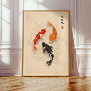 May include: A framed print featuring three koi fish in a pond. One fish is red and white, another is orange, and the third is black and white. The artwork has Japanese calligraphy and a red seal in the upper right corner. The frame is a light brown color.