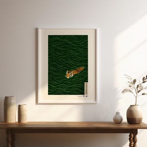 May include: Framed art print featuring a tiger swimming in green water. The tiger is orange and black with a white face. The water has a wavy pattern. The print is in a white frame and is displayed on a wooden table.