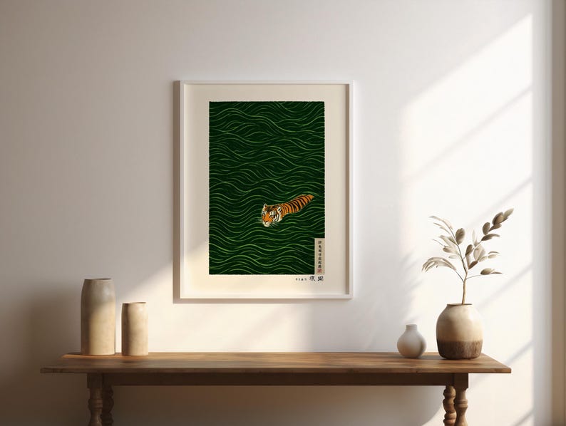 May include: Framed art print featuring a tiger swimming in green water. The tiger is orange and black with a white face. The water has a wavy pattern. The print is in a white frame and is displayed on a wooden table.