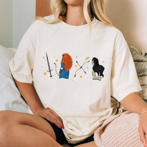 May include: Cream-colored t-shirt featuring a cartoon illustration of a sword, a character with red hair and a blue dress, a bow and arrow, and a black horse. The design includes yellow stars and is printed on a soft, oversized tee.