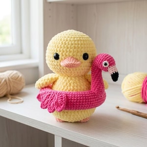 Könnte beinhalten: Eine handgefertigte gelbe Häkelente mit schwarzen Augen und einem rosa Schnabel, die einen rosa Flamingo-Schwimmring trägt. Dieses weiche Spielzeug ist ideal für Kinder oder als Dekorationsartikel. Der Flamingo-Schwimmring verleiht dem Design eine spielerische Note.