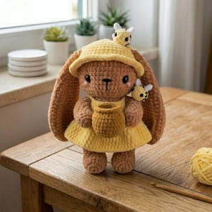 Puede incluir: Un peluche de conejo tejido a ganchillo de color marrón que lleva un sombrero amarillo y un vestido amarillo a juego. El conejo sostiene un pequeño tarro de miel amarillo en sus manos. Dos pequeñas abejas tejidas están unidas al conejo.