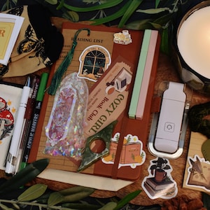 May include: A cozy collection of items including a candle, notebook, stickers, pens, and a small bag with a mushroom design. The items are arranged on a wooden surface, creating a warm and inviting aesthetic. The candle has three lit wicks.