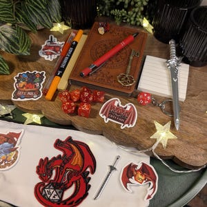 May include: A selection of fantasy role-playing game accessories on a wooden tray. Items include a journal, dice, a miniature sword, and stickers with dragon designs and the text "Let's Roll". Fairy lights and a white pouch are also visible.