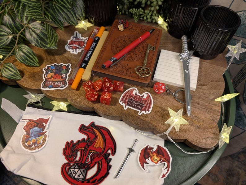 May include: A selection of fantasy role-playing game accessories on a wooden tray. Items include a journal, dice, a miniature sword, and stickers with dragon designs and the text "Let's Roll". Fairy lights and a white pouch are also visible.