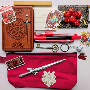 May include: A collection of Dungeons & Dragons themed accessories, including a brown journal with a sword clasp, red pens, dice, stickers, a key, and a red pencil case with a decorative sword. The items are arranged on a white surface.