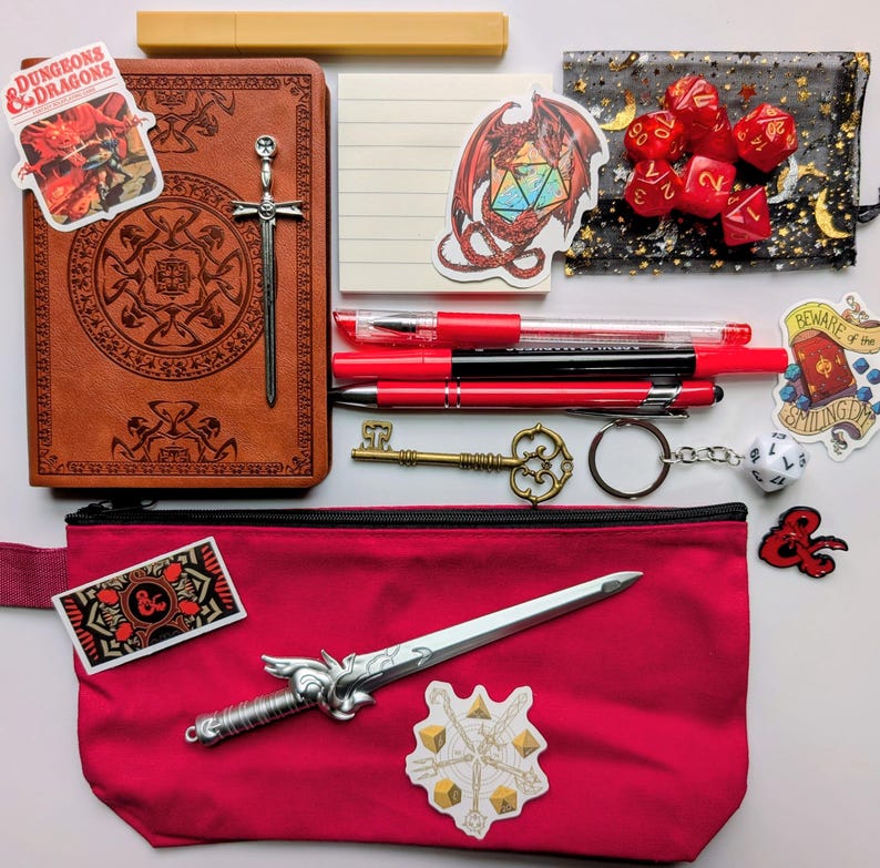 May include: A collection of Dungeons & Dragons themed accessories, including a brown journal with a sword clasp, red pens, dice, stickers, a key, and a red pencil case with a decorative sword. The items are arranged on a white surface.