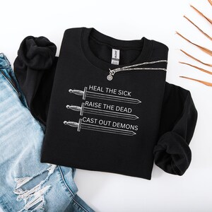 May include: Black crewneck sweatshirt with white text and sword illustrations. The text reads "HEAL THE SICK, RAISE THE DEAD, CAST OUT DEMONS." A silver necklace with a circular pendant is draped across the sweatshirt. The sweatshirt is paired with blue jeans.