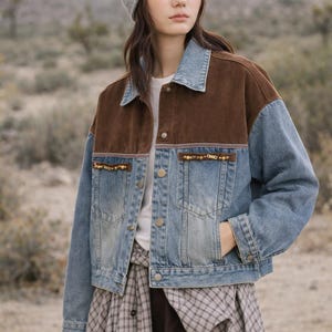 May include: A denim jacket with brown suede-like panels on the shoulders and collar. The jacket has a light blue wash with pockets and button closures. The jacket is decorated with small gold-colored beads. A gray beanie is also visible.