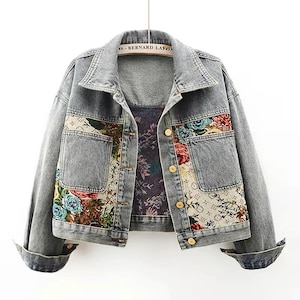 May include: A light gray denim jacket with floral embroidery on the pockets and front panels. The jacket has gold-colored buttons and a patterned interior lining. The sleeves are cuffed at the wrists.