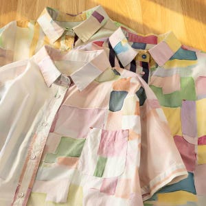 May include: A collection of short-sleeved button-down shirts featuring a multi-colored abstract block pattern in soft pastel shades of pink, yellow, green, and blue. Each shirt has a classic collar, a front button placket, and a single chest pocket.