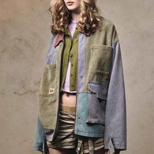 May include: A patchwork jacket in shades of green, blue, and gray, with a corduroy texture. The jacket has multiple pockets and a collared neckline. A green cardigan and a light purple shirt are visible underneath. The outfit is completed with striped shorts.