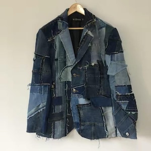 May include: A blazer constructed from various shades of blue denim fabric patches. The garment features a notched lapel, a two-button front closure, and visible frayed edges throughout the seams. The interior is lined with black fabric.