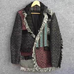 May include: A patchwork blazer with a unique design. The jacket features a mix of textures and colors, including black, gray, and burgundy fabrics. It has fringe detailing along the edges and a patterned panel with green and white stripes.