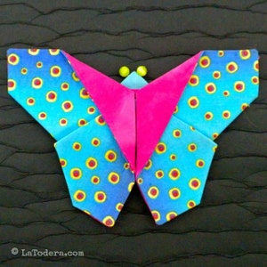 Może przedstawiać: Kolorowy motyl origami z niebieskimi skrzydłami wzorzystymi w żółte i czerwone kółka. Środek motyla jest jasno różowy i ma dwie małe żółte czułki. Origami jest umieszczone na ciemnym, teksturowanym tle.