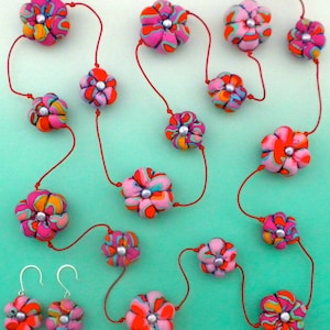 Puede incluir: Un conjunto de joyas florales que incluye un collar y pendientes. El collar y los pendientes están compuestos por cuentas en forma de flor con patrones vibrantes en rosa, naranja y azul, ensartadas en cordones rojos. Cada flor tiene un centro plateado. Los pendientes tienen ganchos plateados.