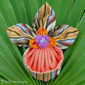 May include: A small, peach-colored fabric pouch with a striped fabric bow. The bow is made of brown, white, and blue stripes and has a large orange and purple flower in the center.